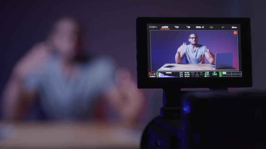A screen where there is a man sitting in front of a laptop and making gestures with his hand while he is talking