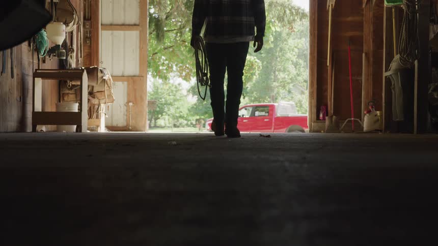 Man walks toward the exit of a rural ranch building carrying a coiled rope