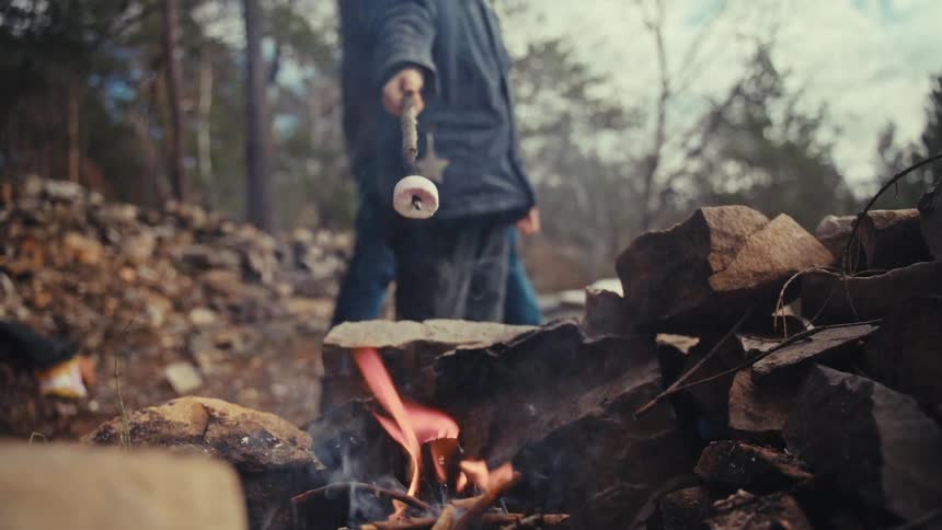 A man wearing a winter jacket holds a piece of food on a stick over a fire to cook while another person walks behind him to pick up a piece of wood at midday
