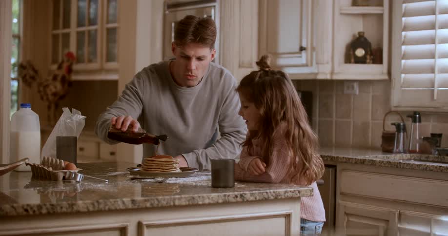 A father pouring syrup on pancakes with his daughter beside him
