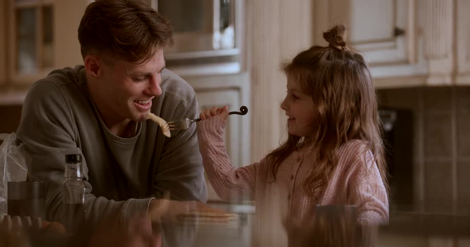 Father and daughter smile while the girl feeds her father as they stand in their kitchen