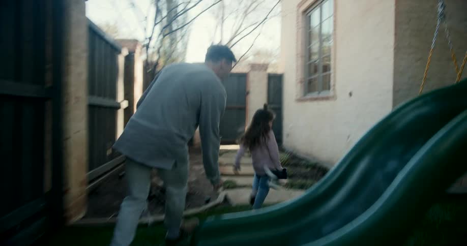 Father chasing his young daughter around wooden swing set in the back yard