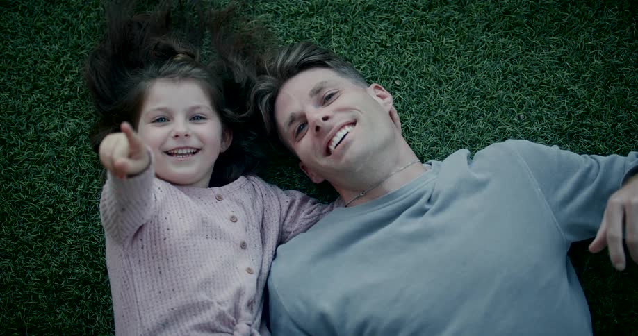 Daughter lying on the turf with her father smiles and points towards the sky