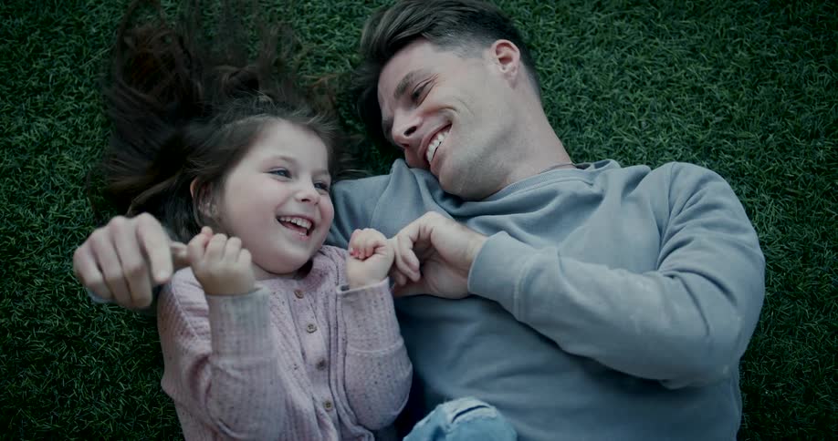 A father and daughter are laying in freshly cut grass laughing and talking