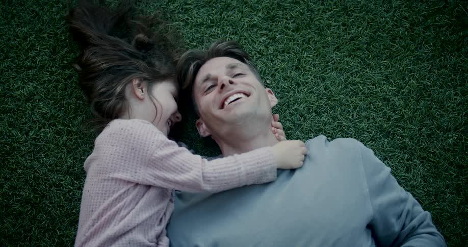 A father and daughter hug and laugh while laying on the grass