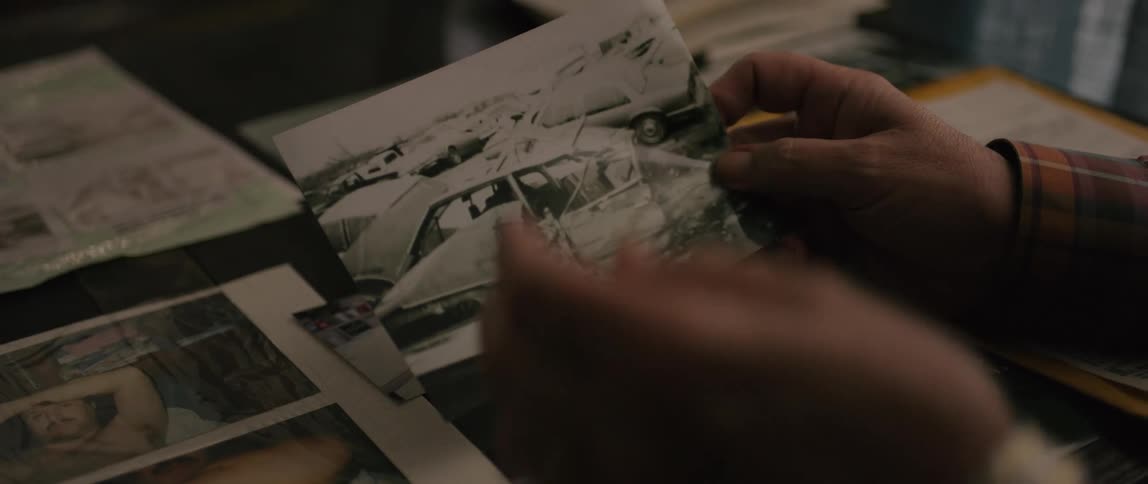 A person holds a blackandwhite photograph of a totaled vehicle above a table covered with albums and photos