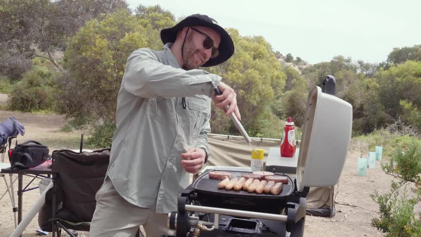 Happy camper grilling hot dogs and burgers on a portable grill while camping in the wilderness