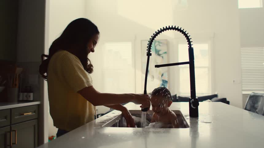 Mom laughing watching her baby splash while taking a bath in kitchen sink