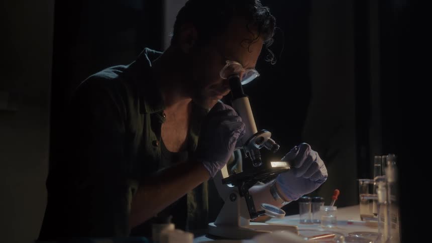 Man wearing gloves examines a sample under a microscope in a dark room