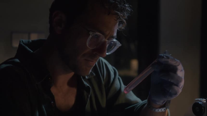 Man examines a vial while sitting in front of a microscope
