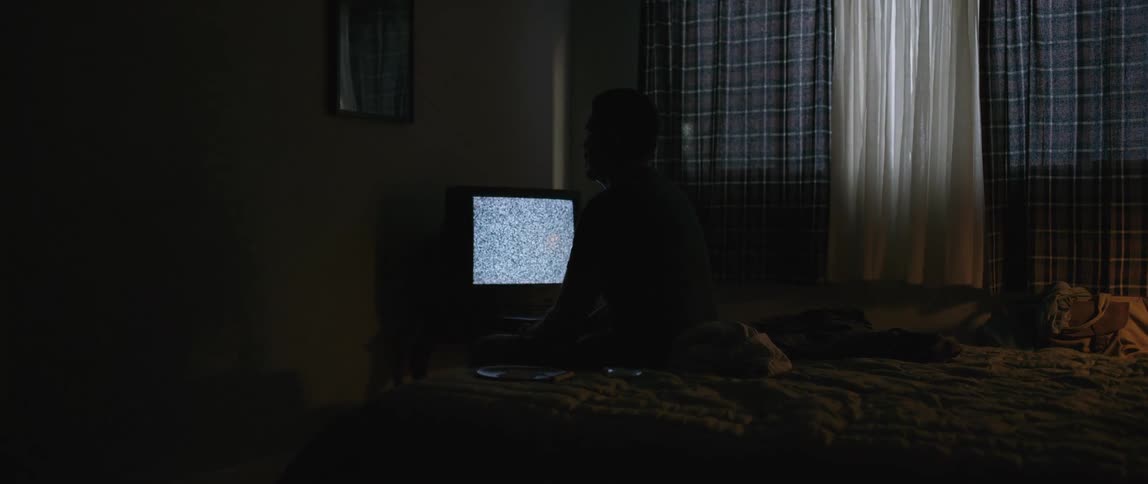 Silhouette of a person sitting on the bed next to a television that is on and has no signal