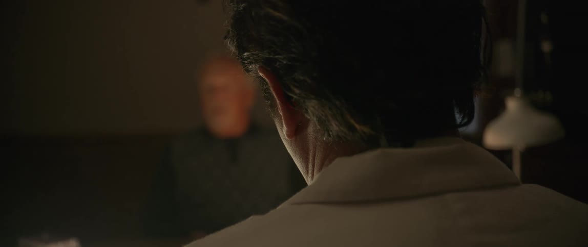 A dimly lit scene showing the back of a mans head as he faces a blurred figure suggesting a tense or private conversation