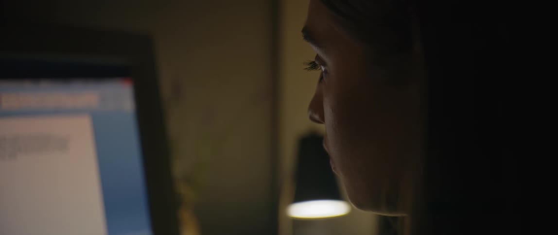 A woman sits in a dimly lit room focused on a computer screen possibly working late at night