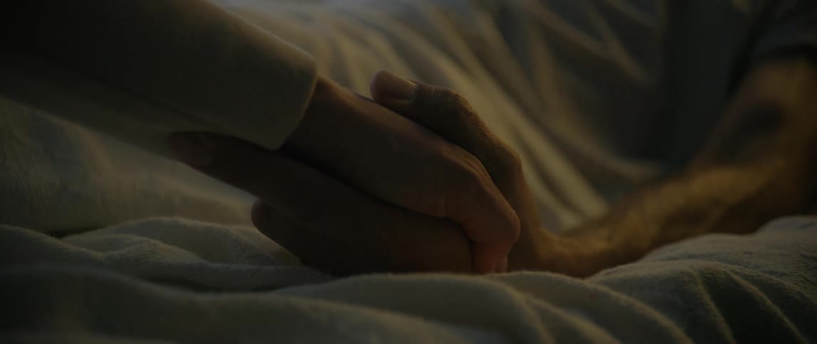 The hands of a sick man in bed showing pain and despair as another person holds his hand