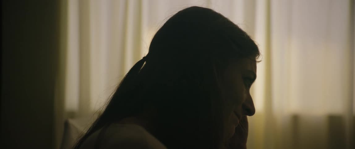 A womans face is partially dark and she is talking to someone who is in front of her and behind her there is a white curtain