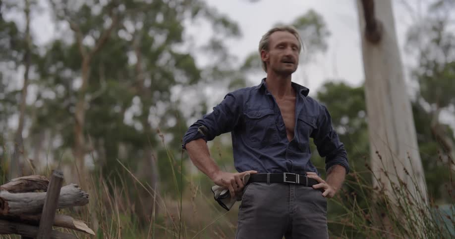 A rugged man stands outdoors in a forest clearing wearing work clothes and holding gloves appearing thoughtful or reflective