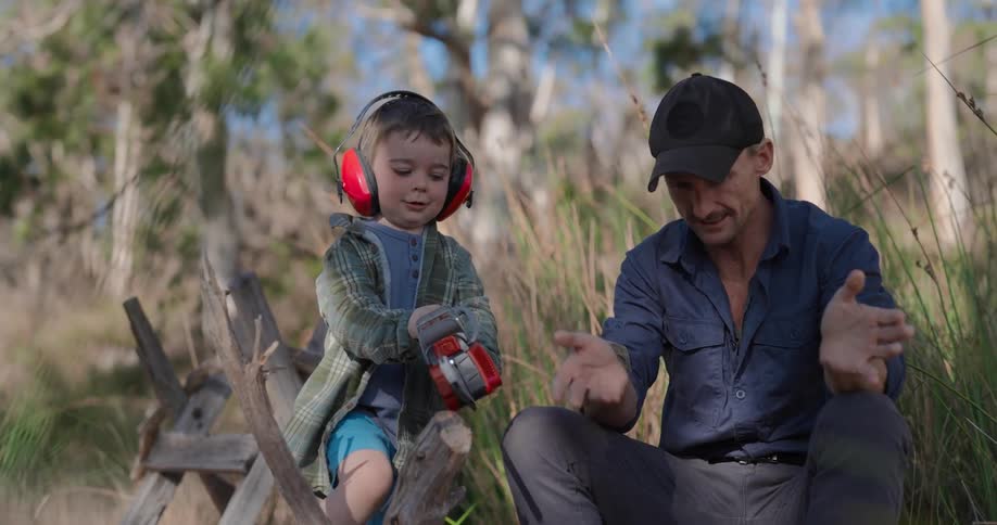 A child is playing with a toy chainsaw as his father holds a branch
