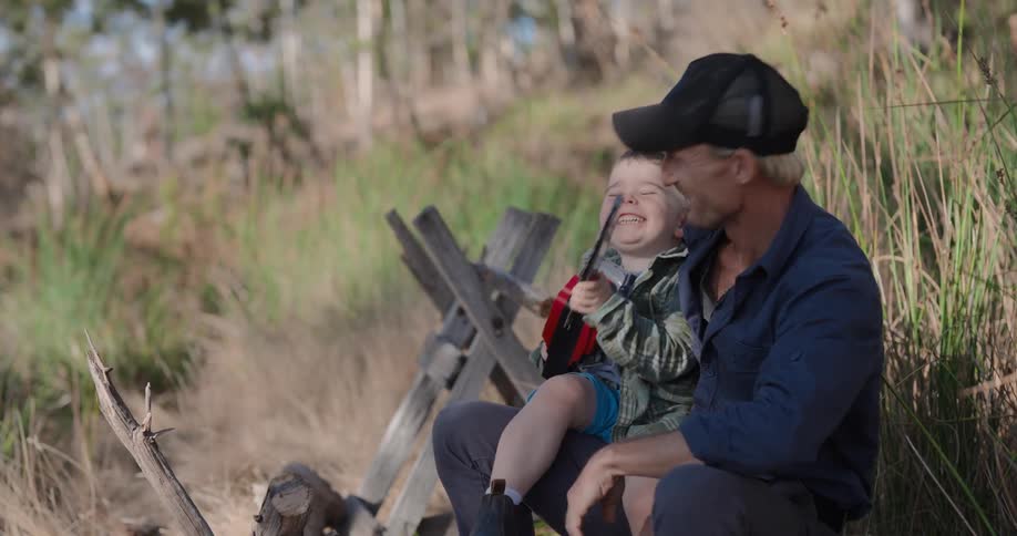 A father and son are sitting outdoors with the boy holding a toy chainsaw and beaming with a smile