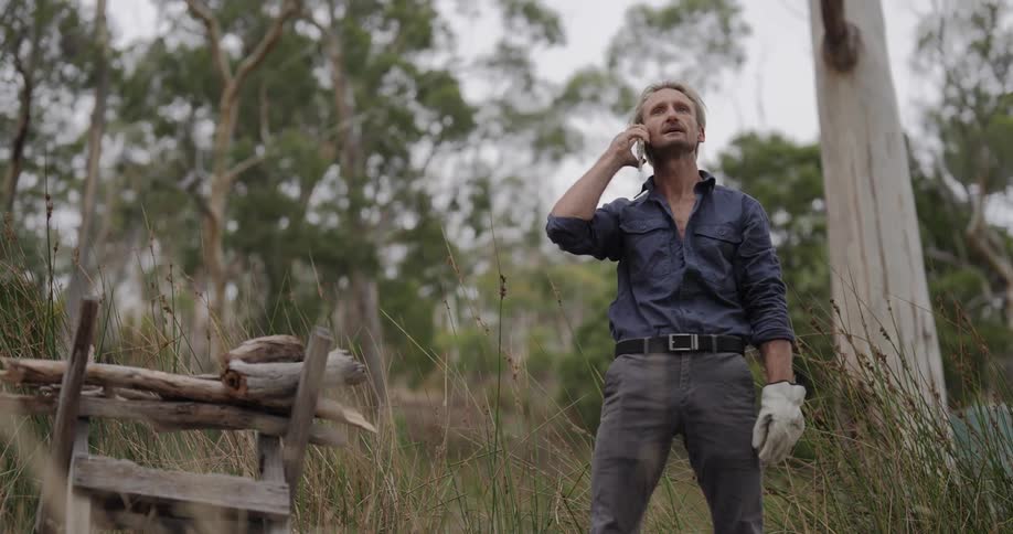 A man stands outdoors in tall grass wearing gloves and talking on a mobile phone appearing to work or inspect something