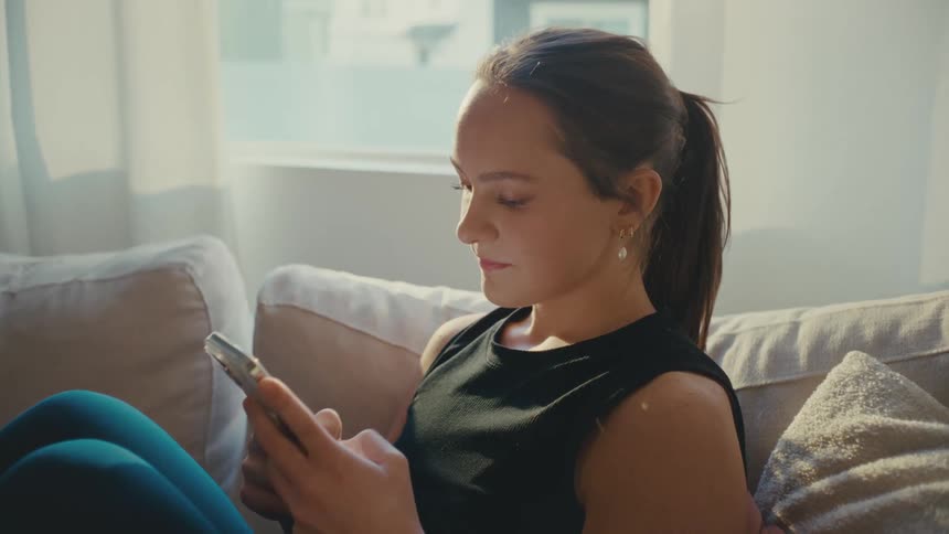 Woman sits on her couch and texts on her smartphone during the day