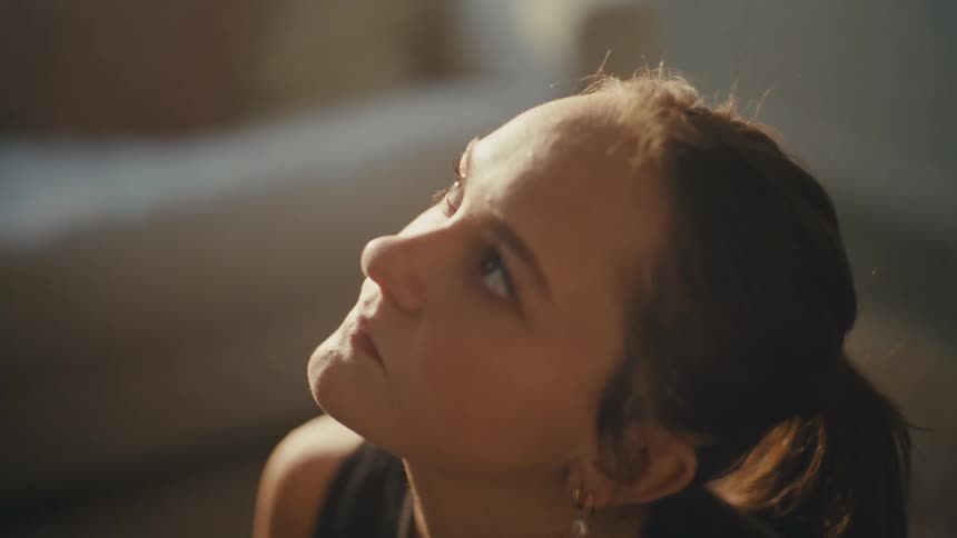 A young woman with a contemplative expression gazes upwards bathed in a warm directional light against a dark background