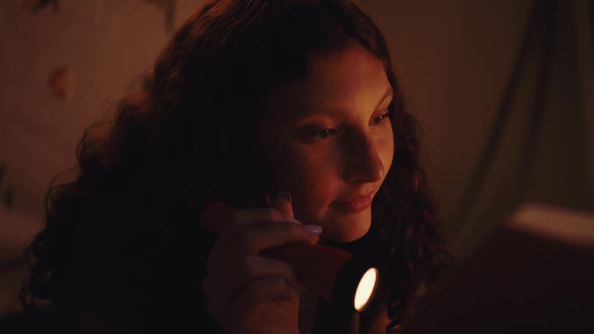 A young girl with curly hair holds a flashlight close to her face softly illuminated as she looks at something offscreen in a cozy dimly lit room