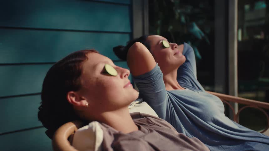 Two women sitting in a yard with cucumbers on their eyes