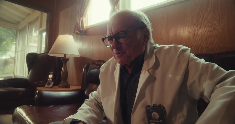 An elderly man wearing a white lab coat passionately gestures while speaking in a warmly lit living room