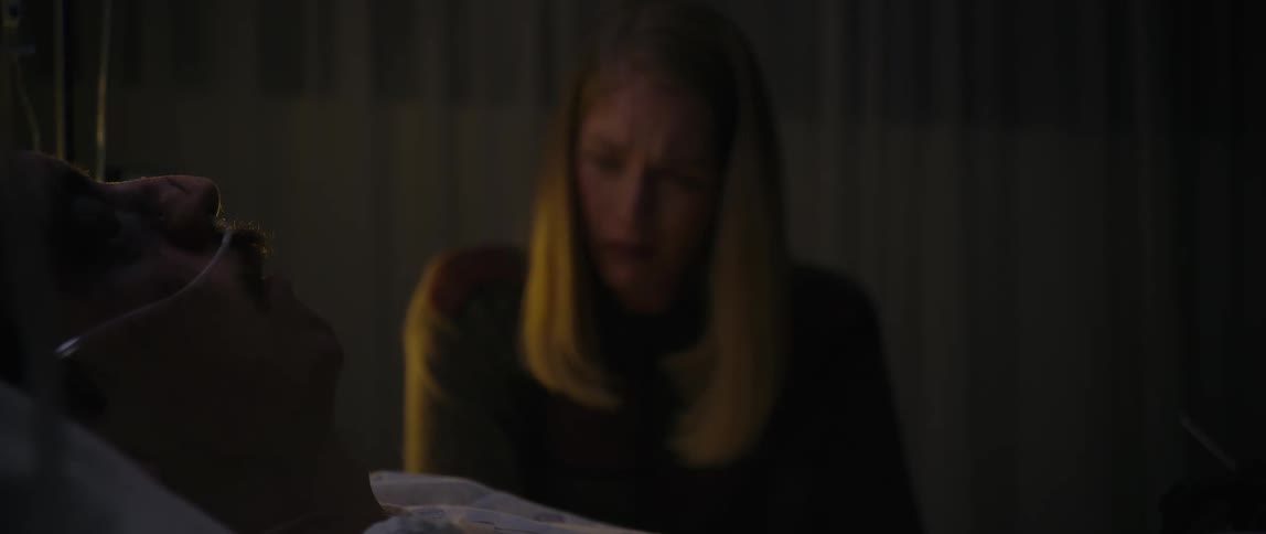 The face of a young blonde woman who is crying and she is looking at a man who is lying in a hospital chamber with an oxygen tube in his nose