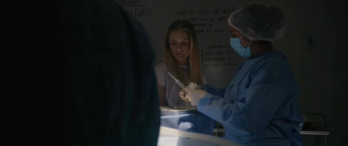 A blonde woman walks around an operation theater as she inspects draped surgeons performing a surgery
