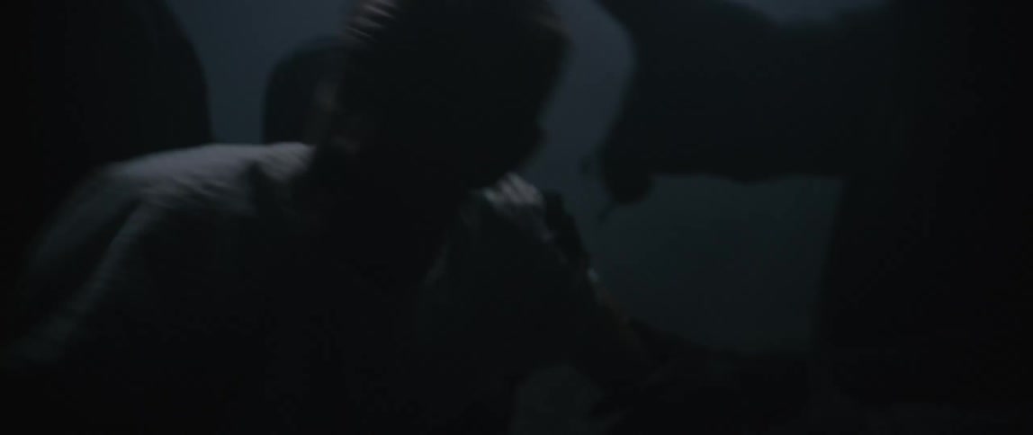Men with tied hair appears among hooded figures in a dark poorly lit environment