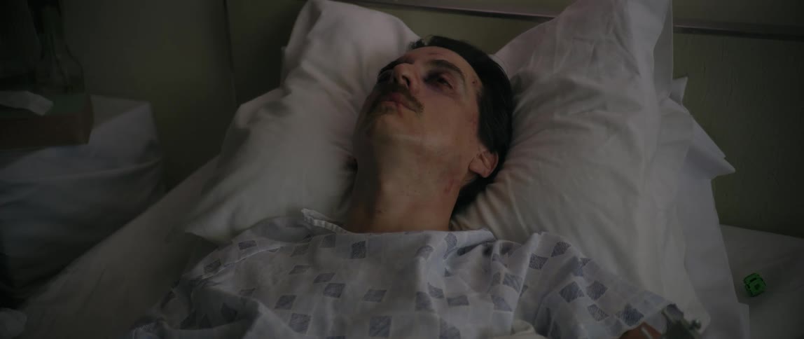 The face of a mustached man who is lying injured in a hospital bed