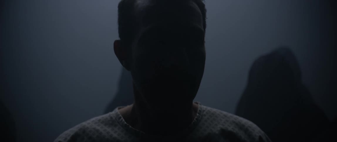 A man turns his head from side to side as he walks in front of a group of people dressed in black in the dark