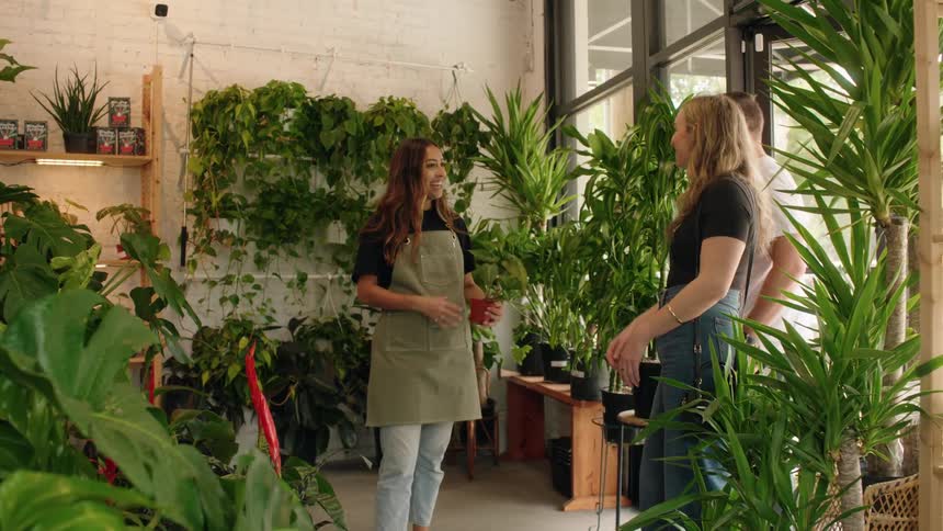 A woman in an apron speaks with two customers inside a lush plantfilled shop