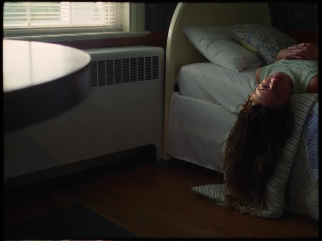 A young woman lies upside down on a bed in a dimly lit room appearing deep in thought or contemplation