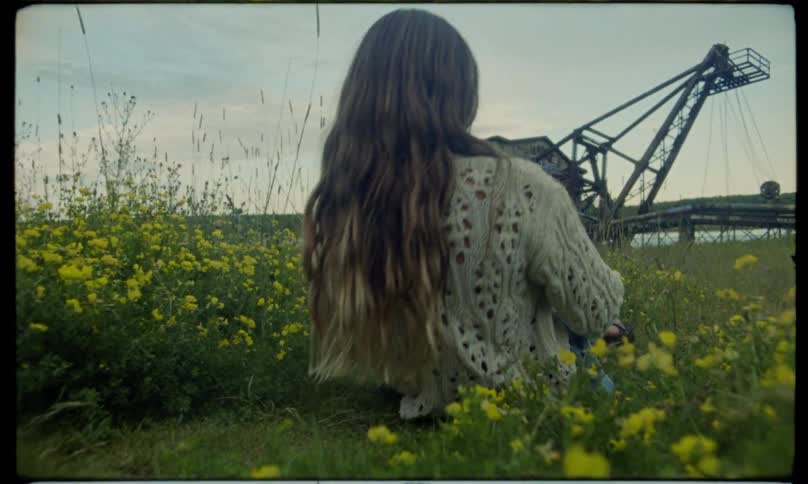 A young woman lies and then sits among yellow wildflowers in front of an abandoned industrial structure reflecting in a quiet natural setting