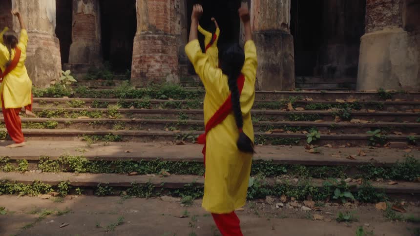 Female dancers in yellow and red traditional attire perform outside an ancient stone structure with columns and steps overgrown with greenery