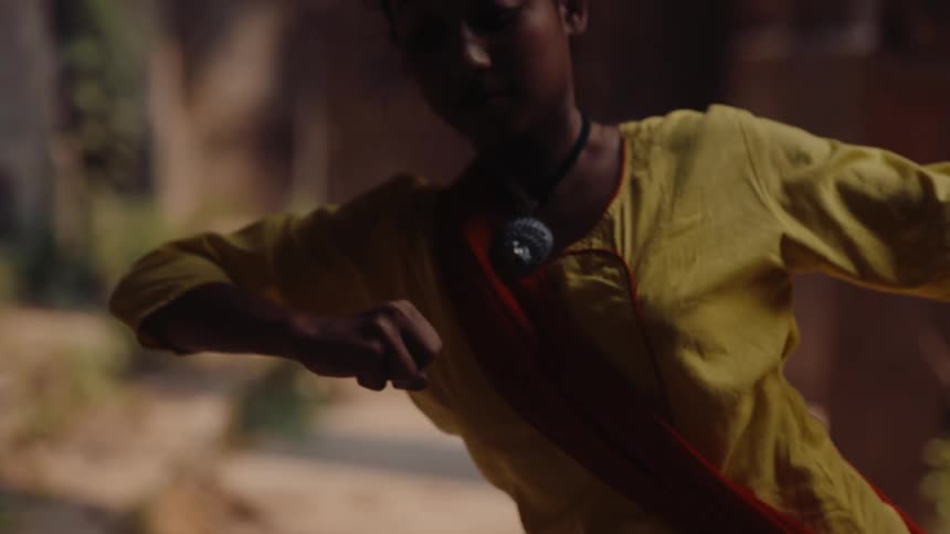 Female dancer wearing a bright tunic moves through dance poses while in the shadow of a colonnade
