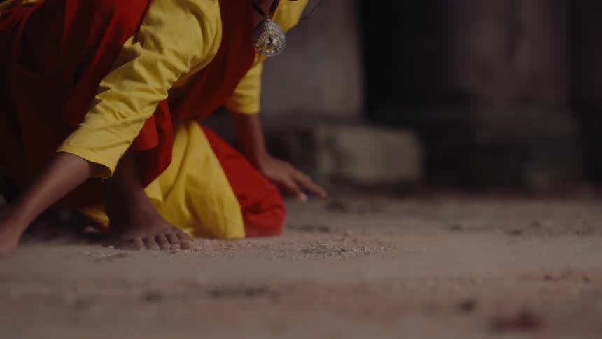 A person dressed in colorful clothing kneels and gently brushes sand or dirt on the ground in a dimly lit environment
