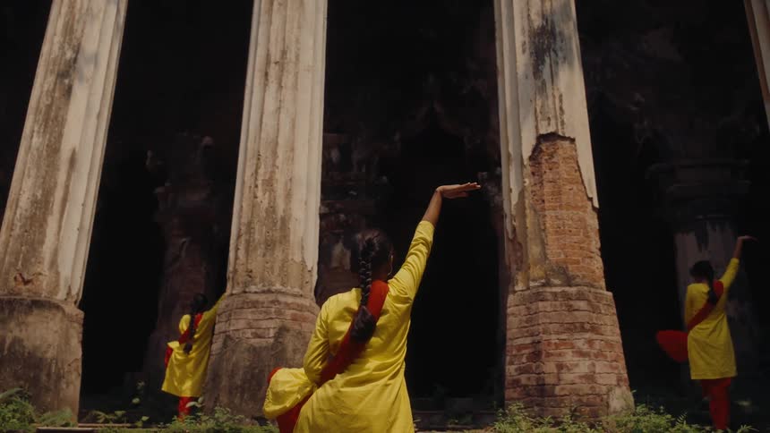 Three women dressed in traditional clothing perform a synchronized classical dance pose in front of ancient stone pillars