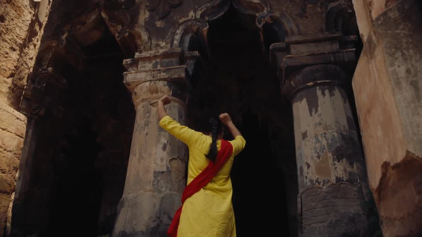 A woman in traditional clothing performs a graceful dance inside an ancient stone structure raising her arms expressively toward the towering pillars
