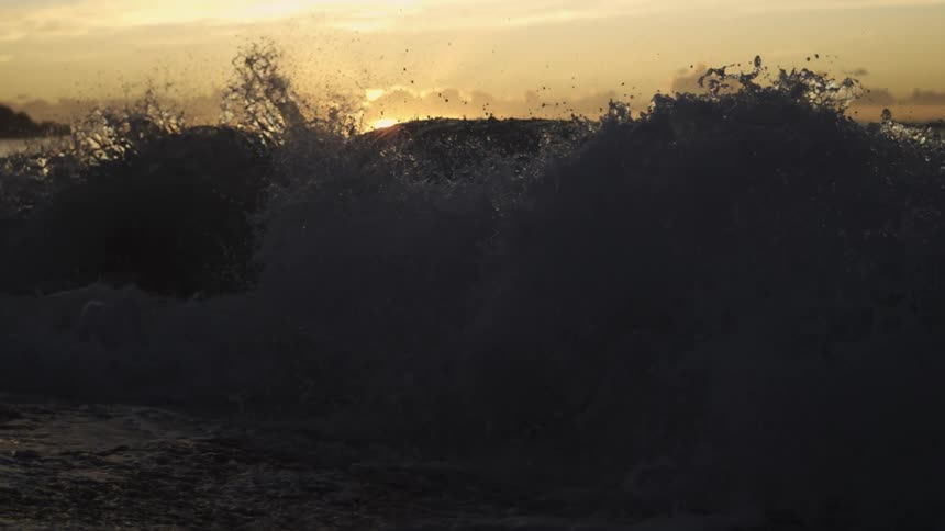 A wave forms and splashes across the ocean surface at dusk