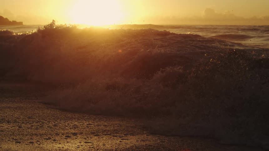 Sun shines from the horizon onto a wave crashing onto the beach