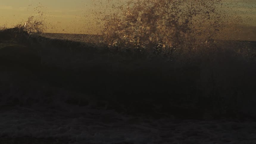 A sea wave breaking slowly and throwing several drops of water upwards on a morning with an orange sky