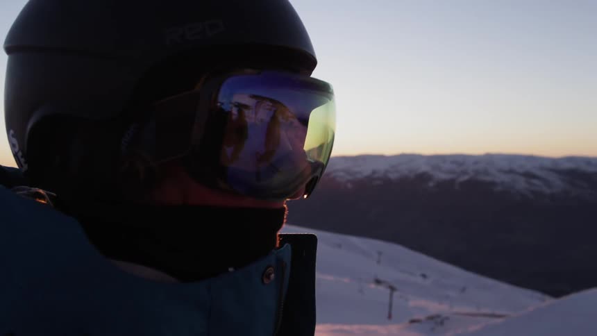 Skier wearing helmet stands atop snow covered mountain at sunset
