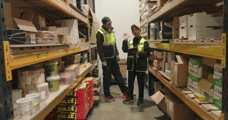 Two warehouse workers in reflective gear discuss inventory in a packed storage aisle