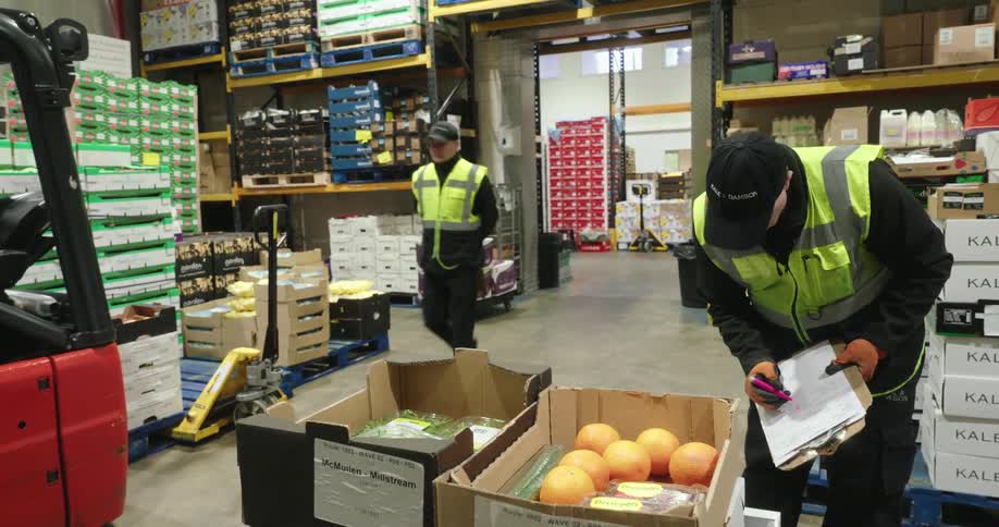 Workers in high visibility vests are checking inventory among pallets and boxes of produce in a warehouse