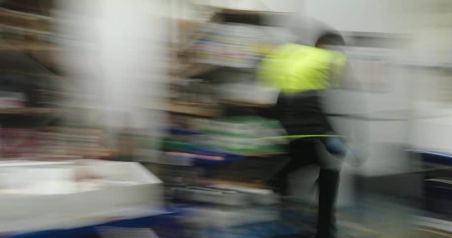 A man wearing a safety vest rolls a cart and restocks items on a shelf inside a warehouse