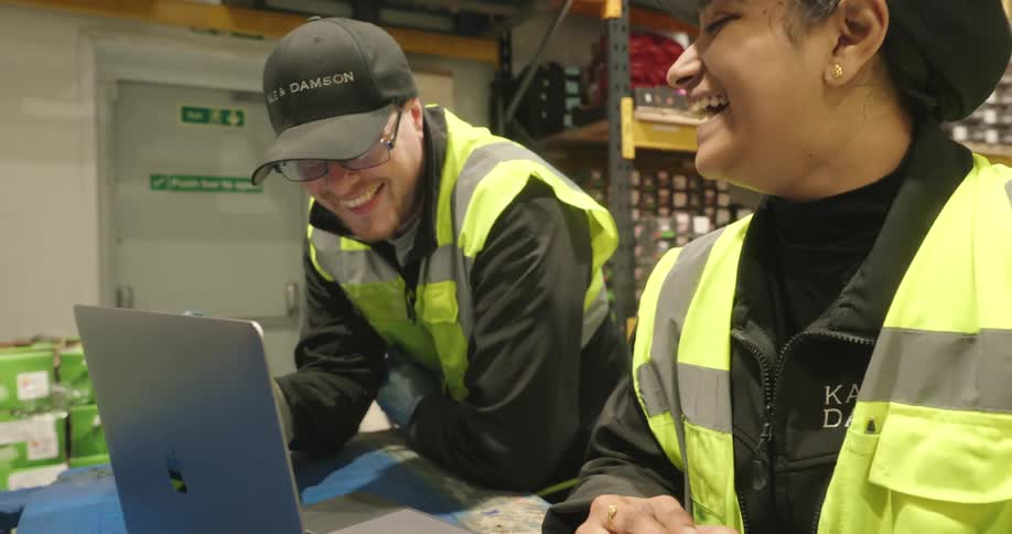 Man and woman talks and laughs while standing in a warehouse with an opened laptop