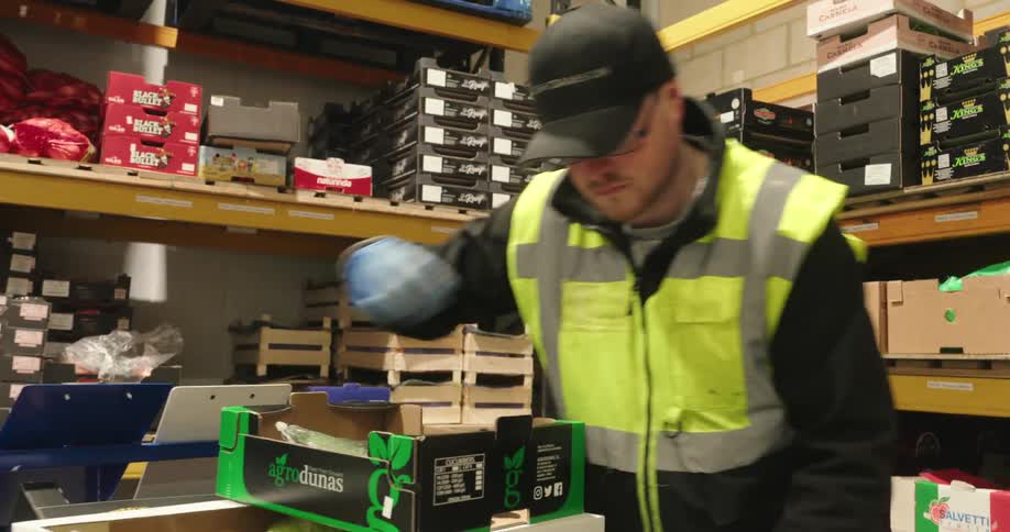 A worker wearing a high visibility vest and gloves is packing boxes labeled kale damson in a well stocked warehouse